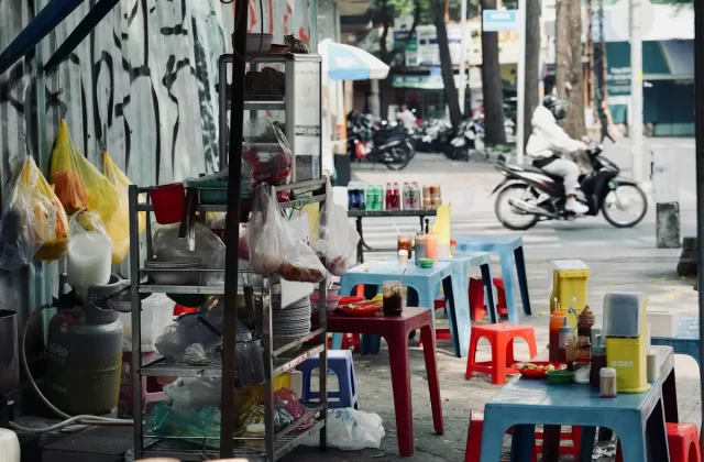 Quán ăn vỉa hè với bàn ghế nhựa dưới tán dù, xe máy chạy ngang