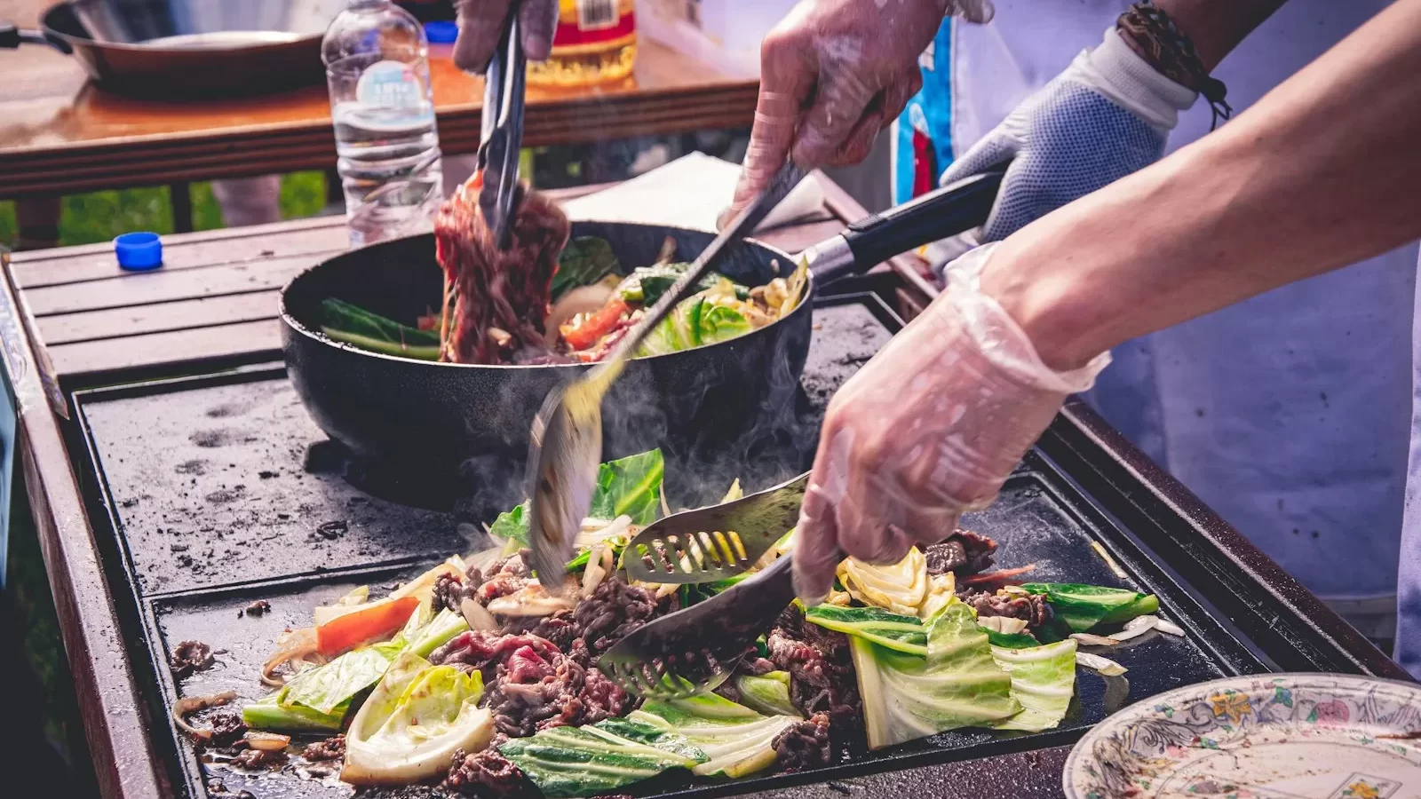 Đôi tay đeo găng đảo thịt và rau cải trong chảo trên bếp lửa lớn