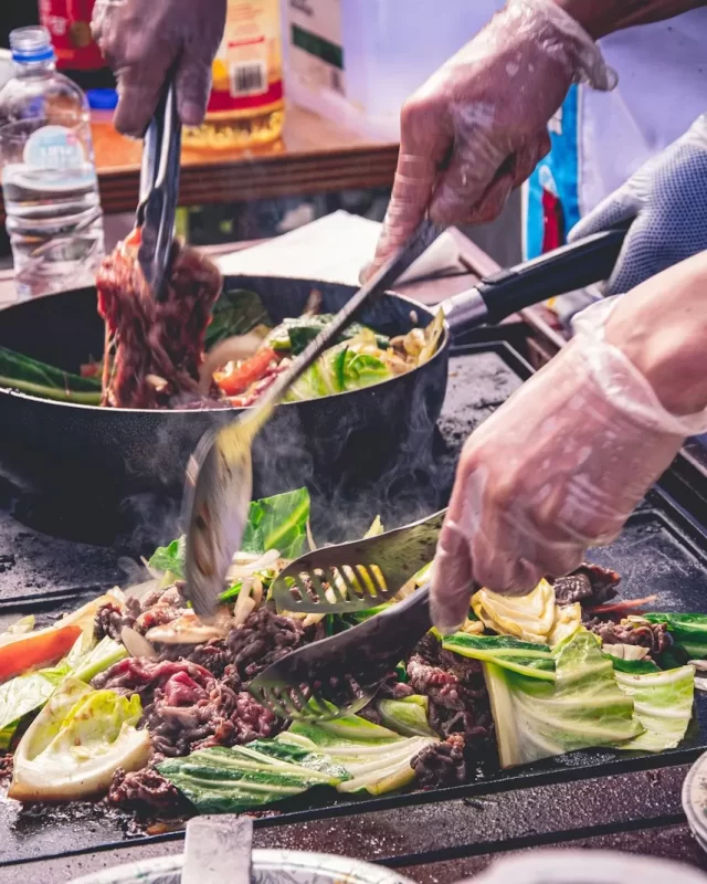 Đôi tay đeo găng đảo thịt và rau cải trong chảo trên bếp lửa lớn