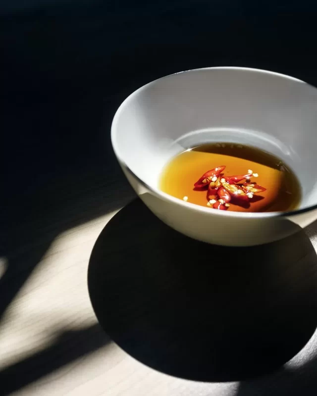 Chén sứ trắng đựng nước mắm màu hổ phách với ớt đỏ thái lát, nắng chiếu nghiêng