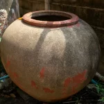 Chum sành lớn miệng loe đặt sát tường gạch, thân chum loang màu thời gian