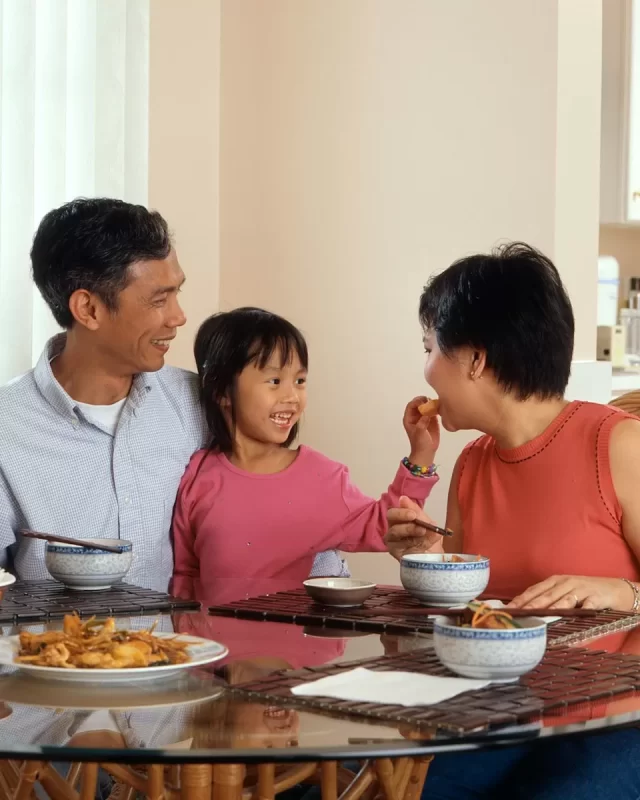 Gia đình ba người quây quần bên bàn ăn, mẹ đút cho con gái miếng ăn