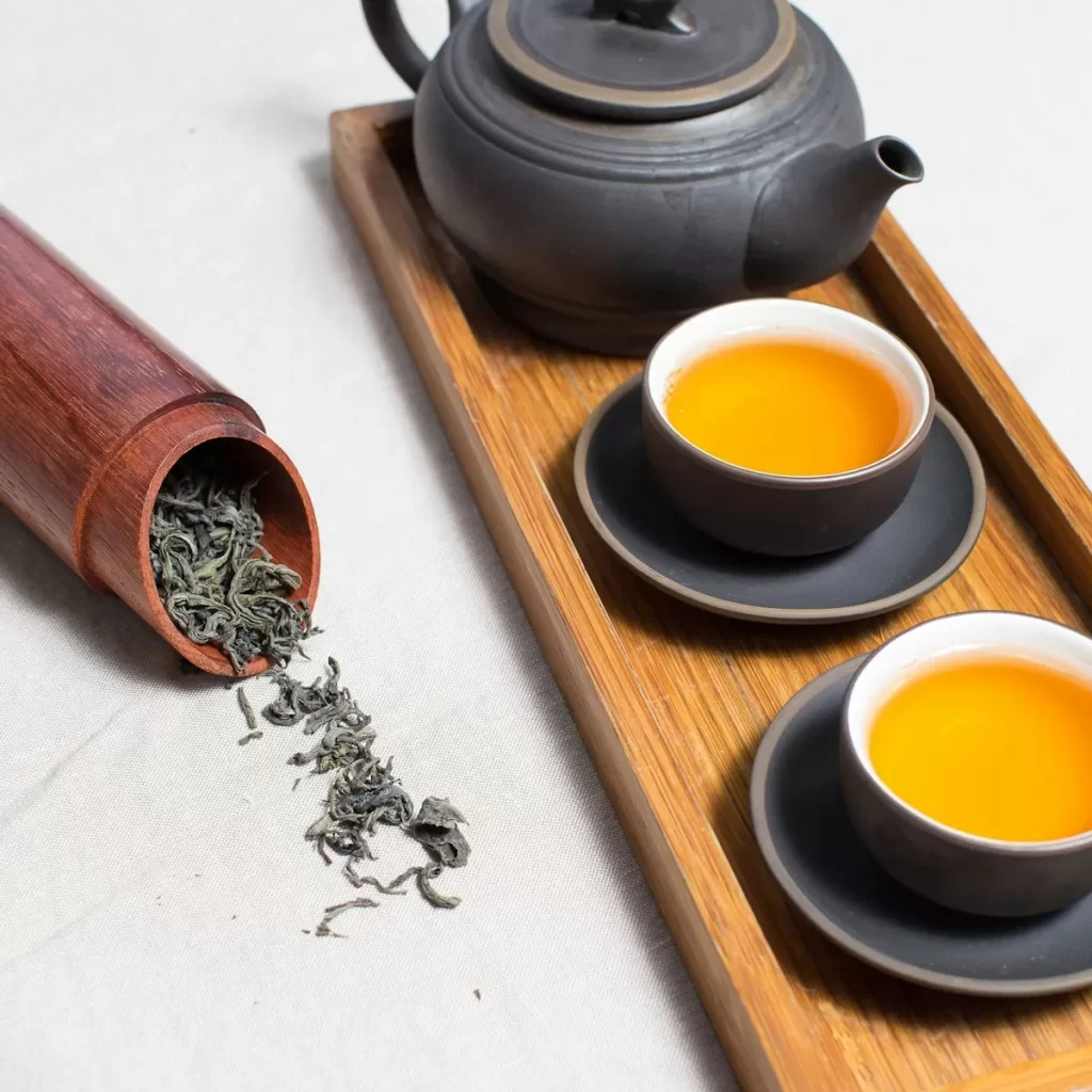 Ấm trà gốm sẫm và hai chén trà vàng sóng sánh trên khay gỗ, ống trà đổ lá khô