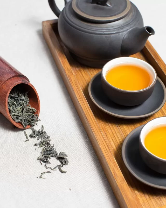 Ấm trà gốm sẫm và hai chén trà vàng sóng sánh trên khay gỗ, ống trà đổ lá khô