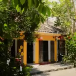 Mặt tiền ngôi nhà vàng có hàng hiên nhỏ và cây xanh trong hẻm Hội An, nơi đặt homestay Góc Nhỏ · Hẻm Cau Lầu