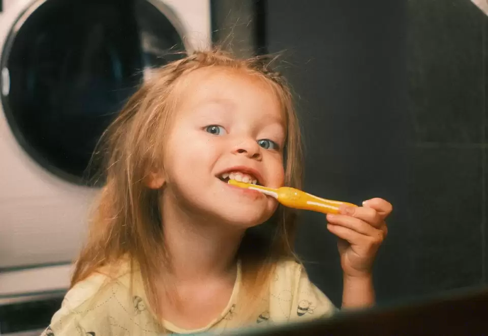 Bé gái vừa đánh răng vừa cười trong phòng tắm, thói quen buổi sáng của bé