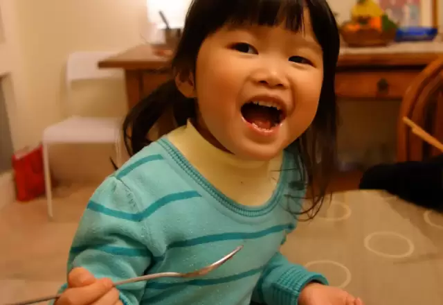 Bé gái cười tươi hết cỡ, tự cầm thìa xúc ăn bên bàn ăn gia đình