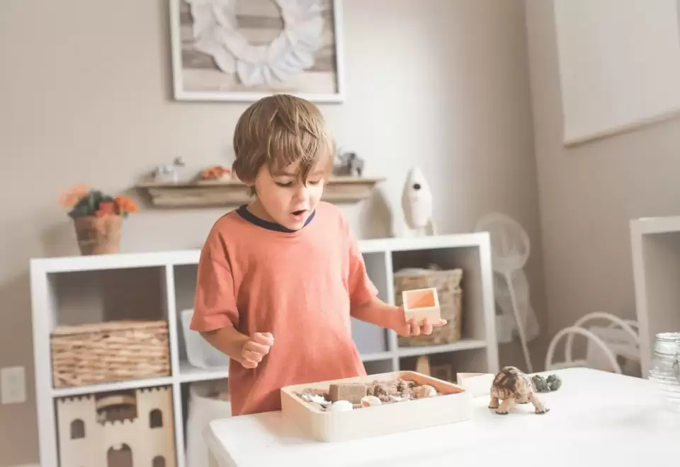 Bé trai tập trung chọn đồ vật trên khay gỗ cạnh kệ thấp trong góc lớp Montessori sáng nhẹ