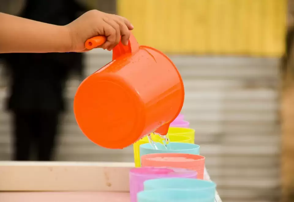 Tay bé rót nước từ chiếc ca cam sang dãy ly nhỏ nhiều màu trên bàn, bài tập thực hành cuộc sống