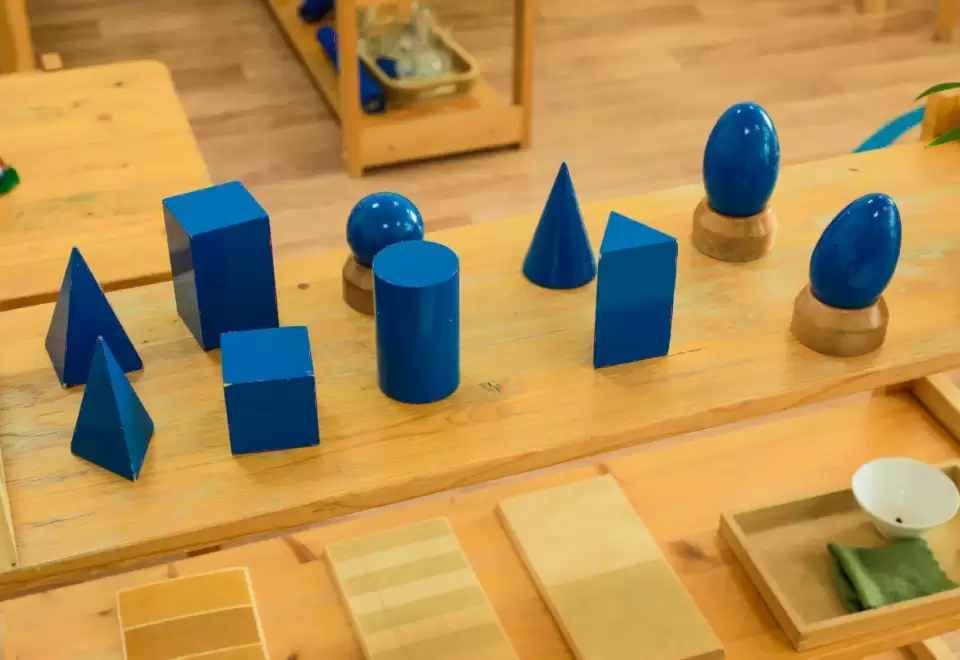 Các khối hình học màu xanh dương gồm trụ, nón, cầu đặt trên thảm giáo cụ gỗ Montessori