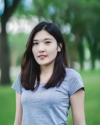 Mẹ bé trẻ tóc dài, áo thun xám xanh, đứng nhìn thẳng trong công viên xanh