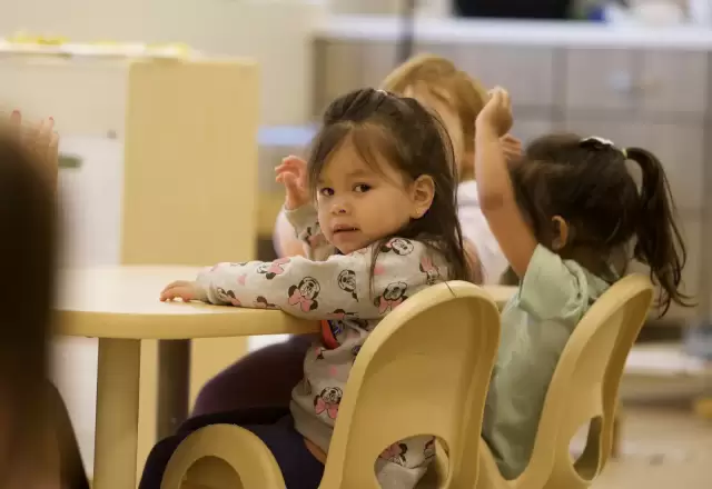 Mấy bé ngồi bên bàn tròn trên ghế nhỏ màu vàng, nhìn quanh lớp học tò mò
