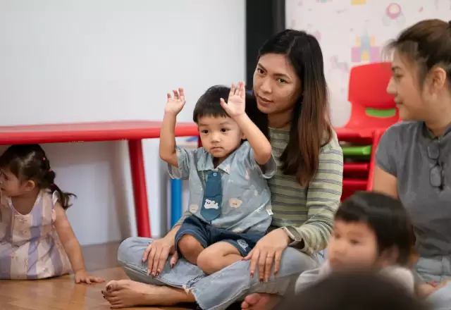 Cô giáo ngồi bệt ôm bé nhỏ giơ hai tay vui vẻ giữa lớp học nhà trẻ
