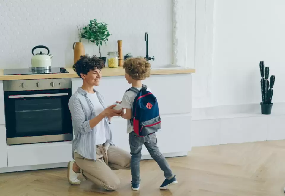 Cô ngồi xổm chào đón bé đeo ba lô ở lối vào, đón bé vào lớp an toàn