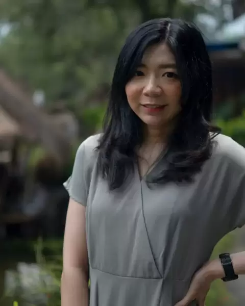 Cô giáo tóc đen dài, áo xám, ánh mắt dịu dàng trước nền cây xanh mờ