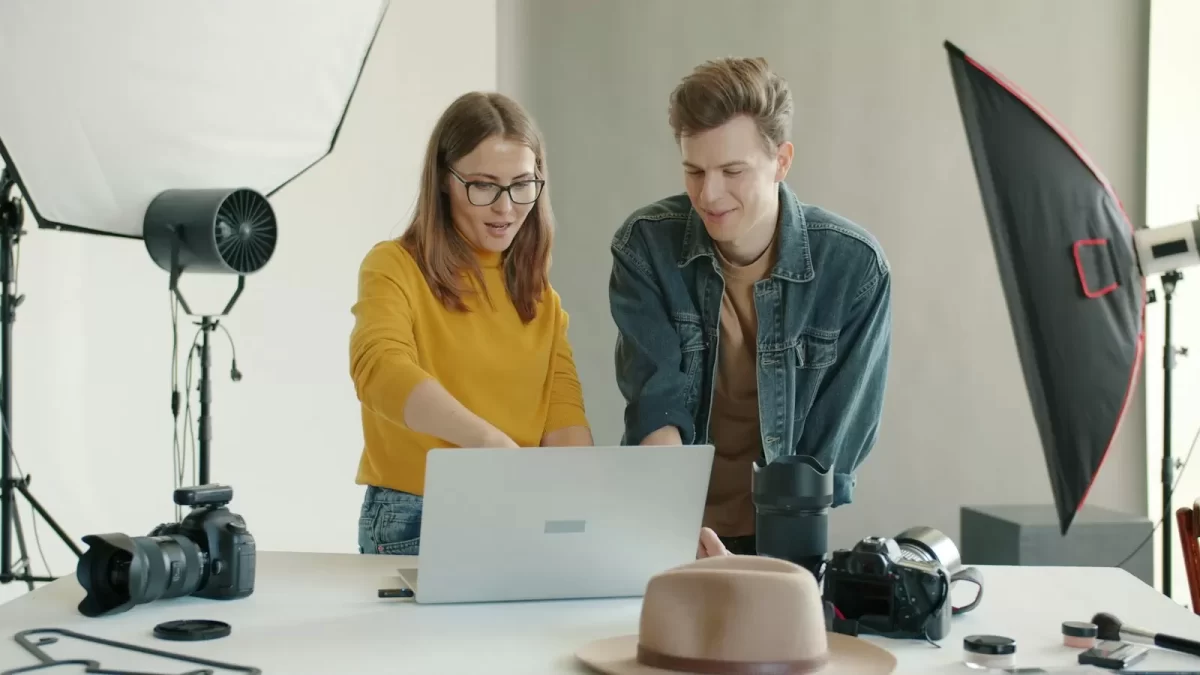 Hai người cùng xem ảnh trên laptop trong studio có đèn softbox và máy ảnh