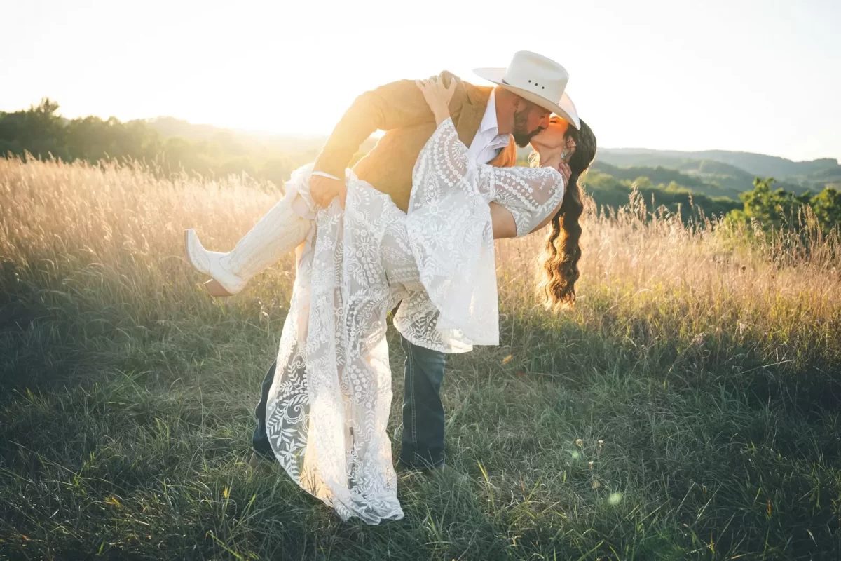 Chú rể nghiêng người hôn cô dâu váy ren trên đồng cỏ vàng