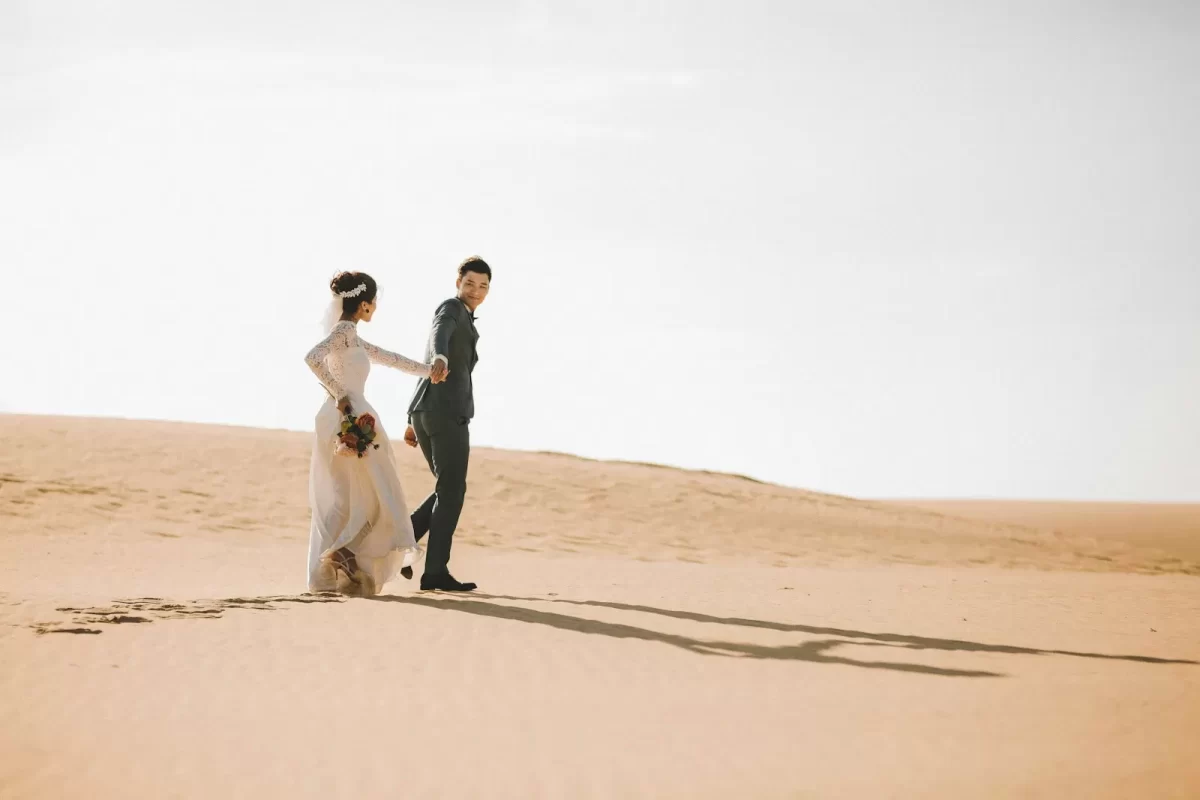 Cô dâu váy trắng và chú rể đứng giữa đồi cát rộng, bầu trời sáng
