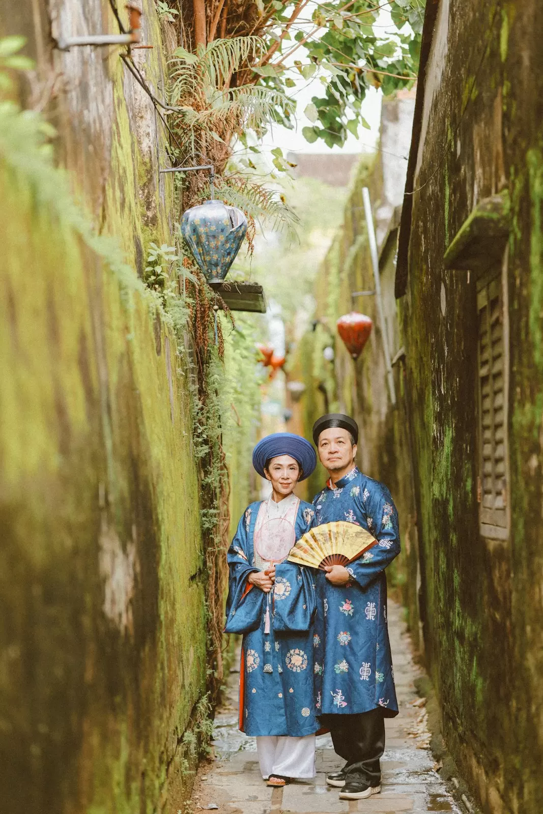 Cặp đôi áo dài ngũ thân xanh đứng trong con hẻm rêu phong, tay cầm quạt