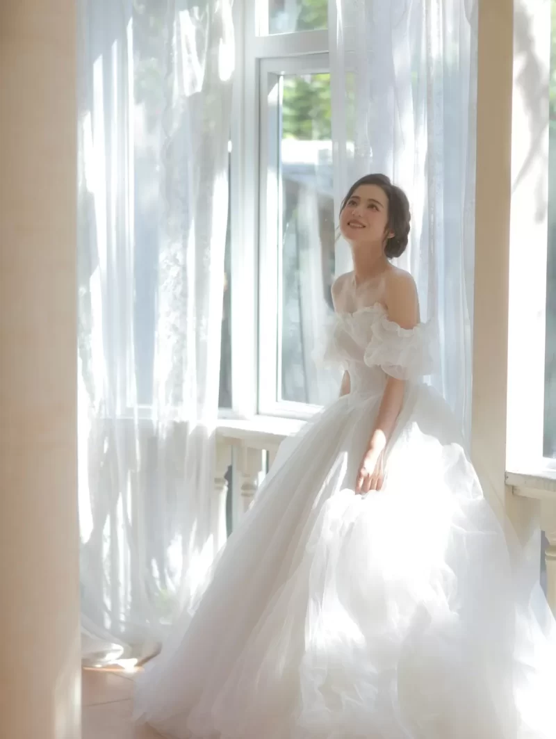 Cô dâu váy tulle xoè nhiều lớp đứng cạnh cửa sổ ngập nắng