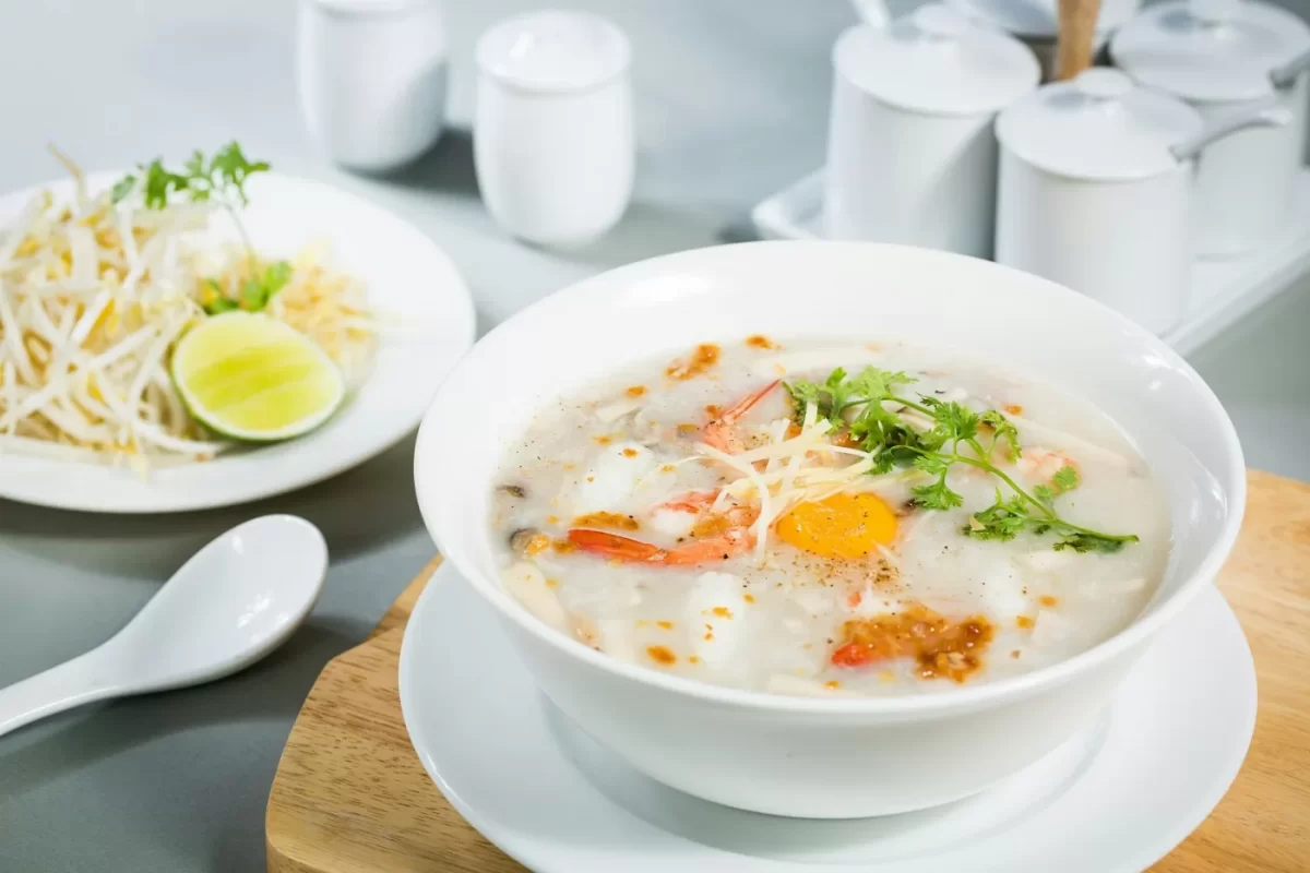 Tô cháo nóng với trứng và rau thơm, đặt cạnh đĩa giá đỗ và chanh