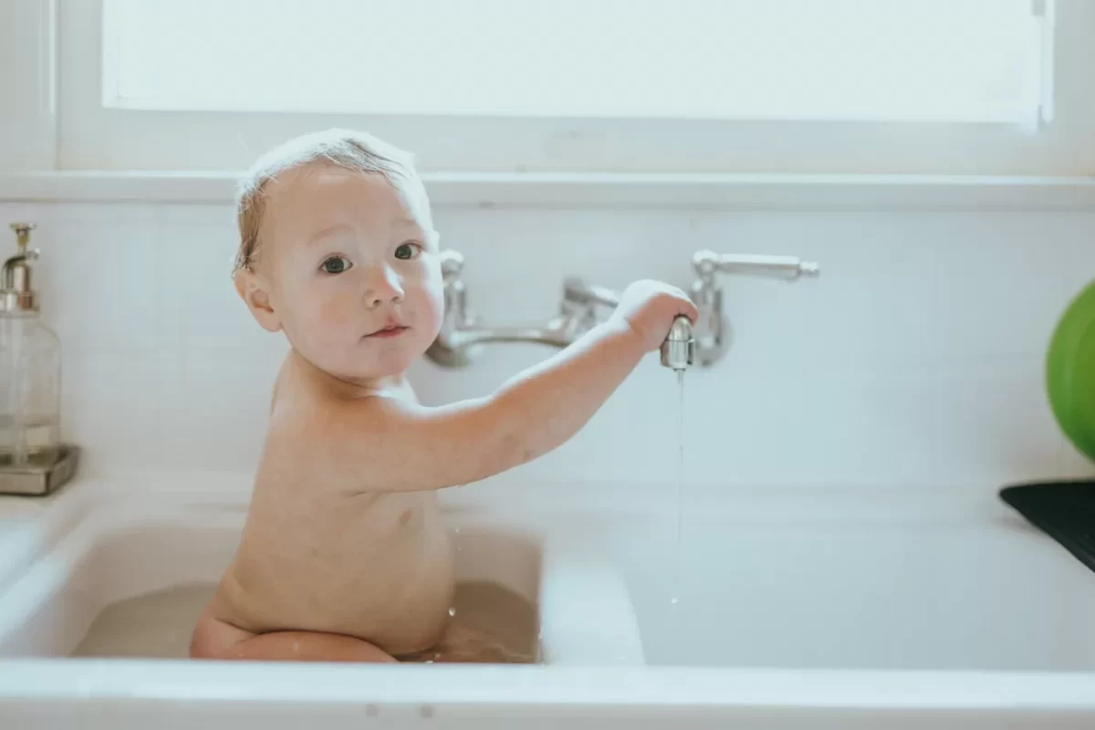 Em bé ngồi trong bồn rửa trắng, với tay chạm vào vòi nước