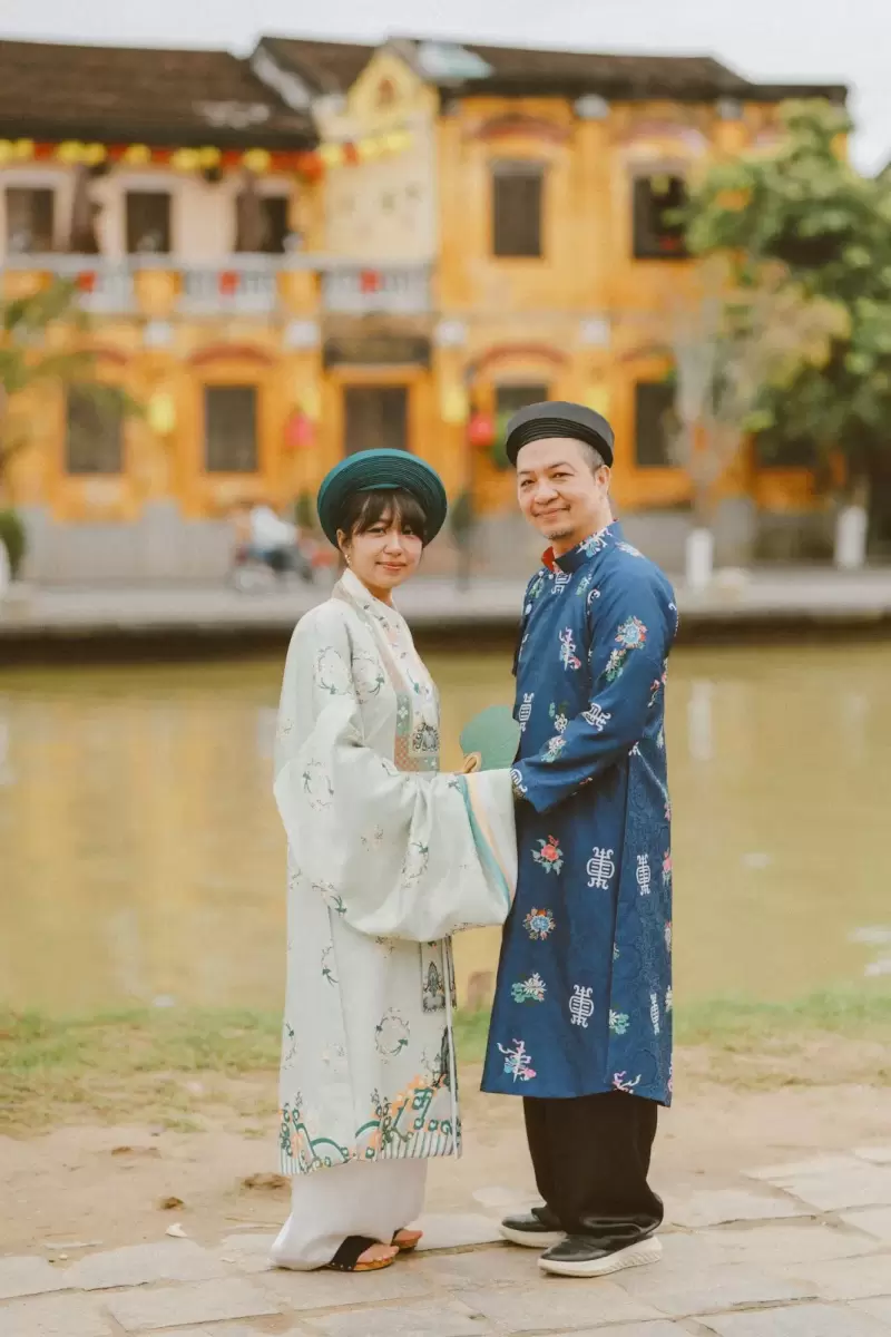 Ảnh cưới Hội An phong cách Cổ điển: cặp đôi áo dài đứng cạnh nhau bên bờ sông, sau lưng là dãy nhà vàng