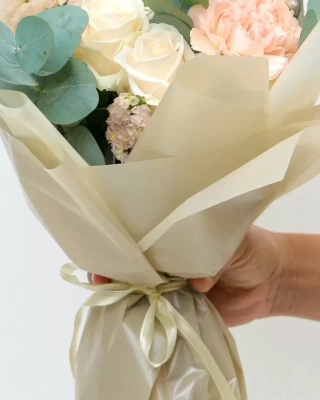 Cận cảnh: bó hoa pastel hồng kem gói giấy, gồm hồng, mẫu đơn và lisianthus