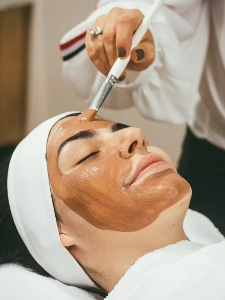 Kỹ thuật viên dùng cọ thoa mặt nạ lên mặt khách, đầu quấn khăn trắng